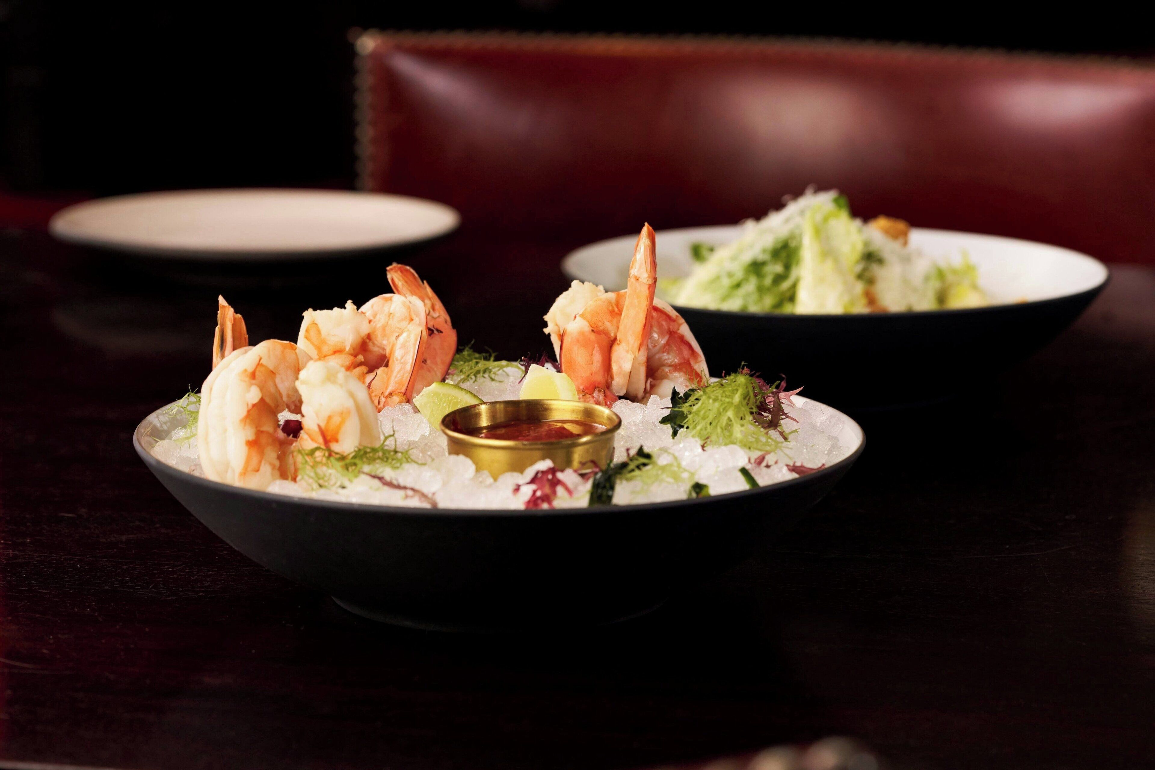 Une table à manger avec une assiette de crevettes et de salade, un saladier rempli de salade et un petit récipient contenant de la sauce.