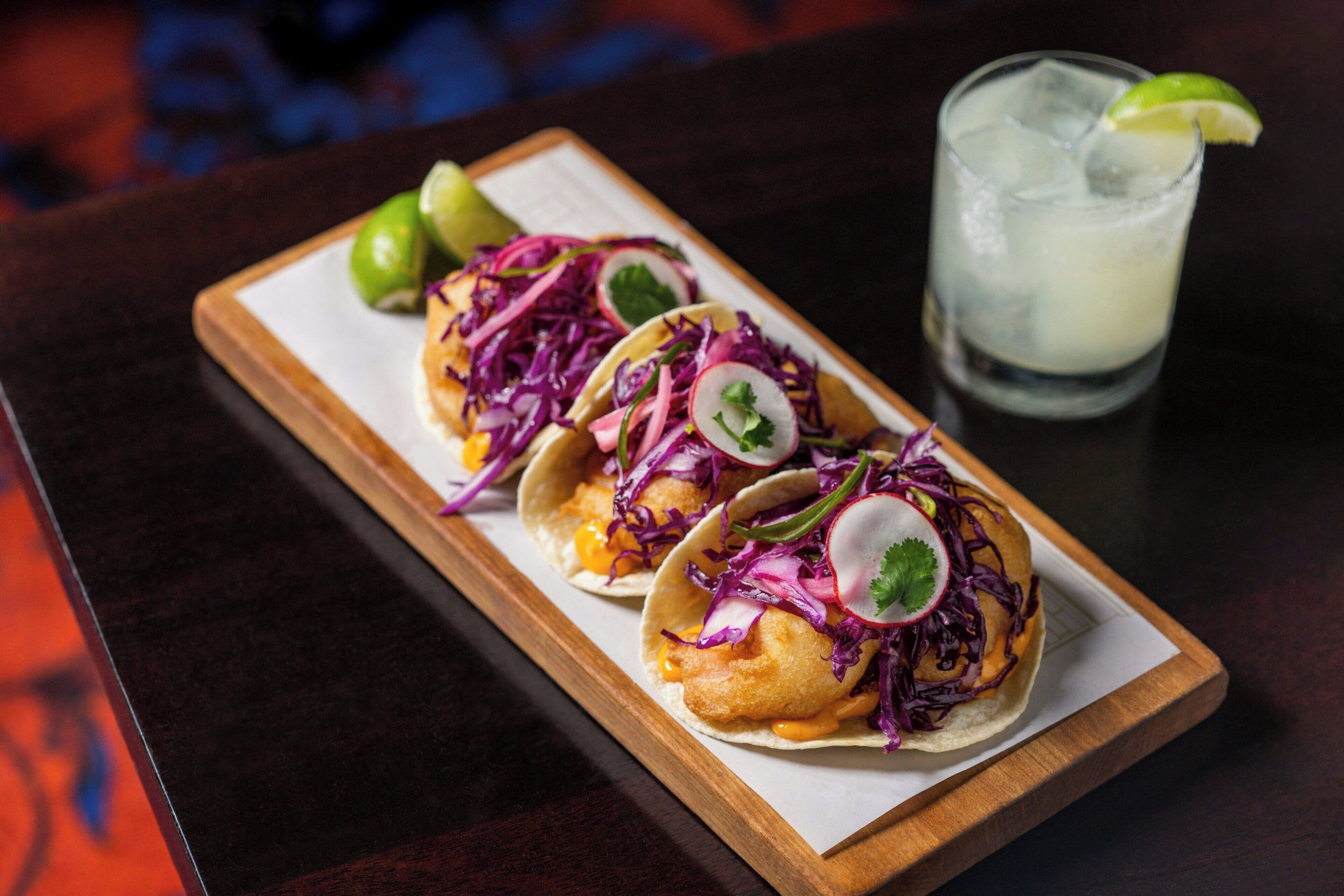 Drei Tacos mit Limettenspalte und einem Glas Eistee auf einem Holztablett.