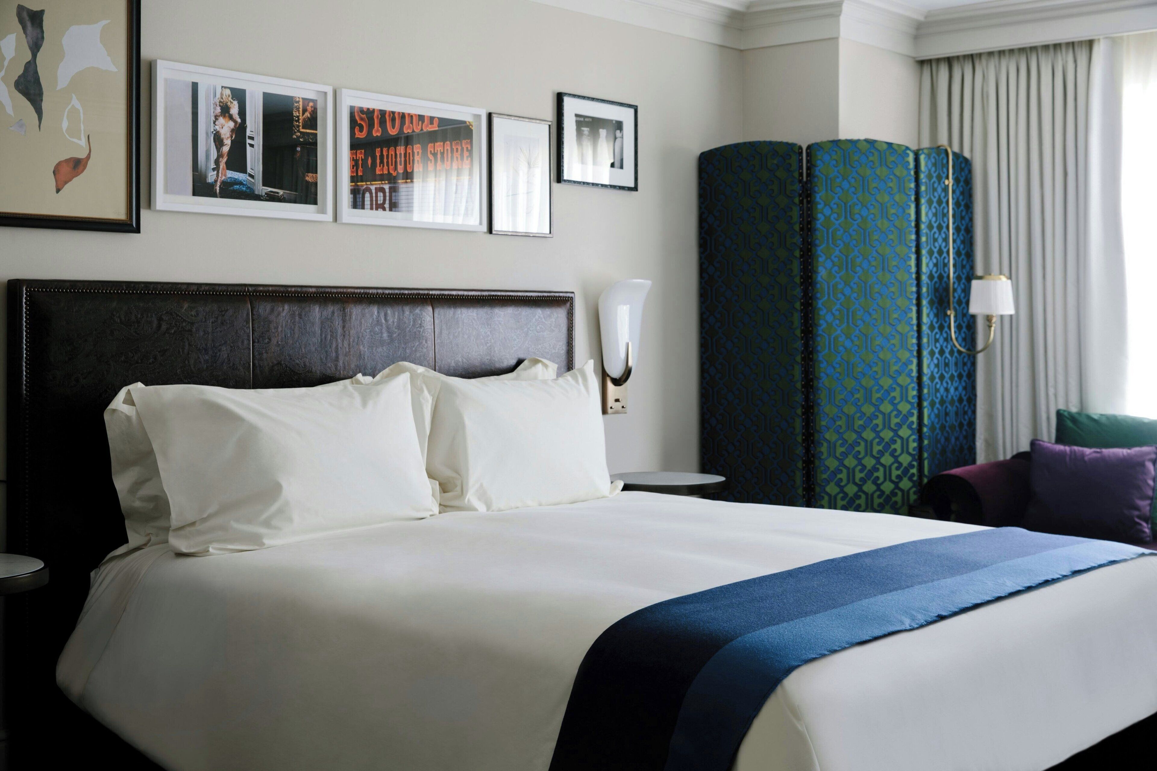 Habitación de hotel con una cama grande, una mesita de noche, una lámpara de pared y un amplio ventanal con cortinas.