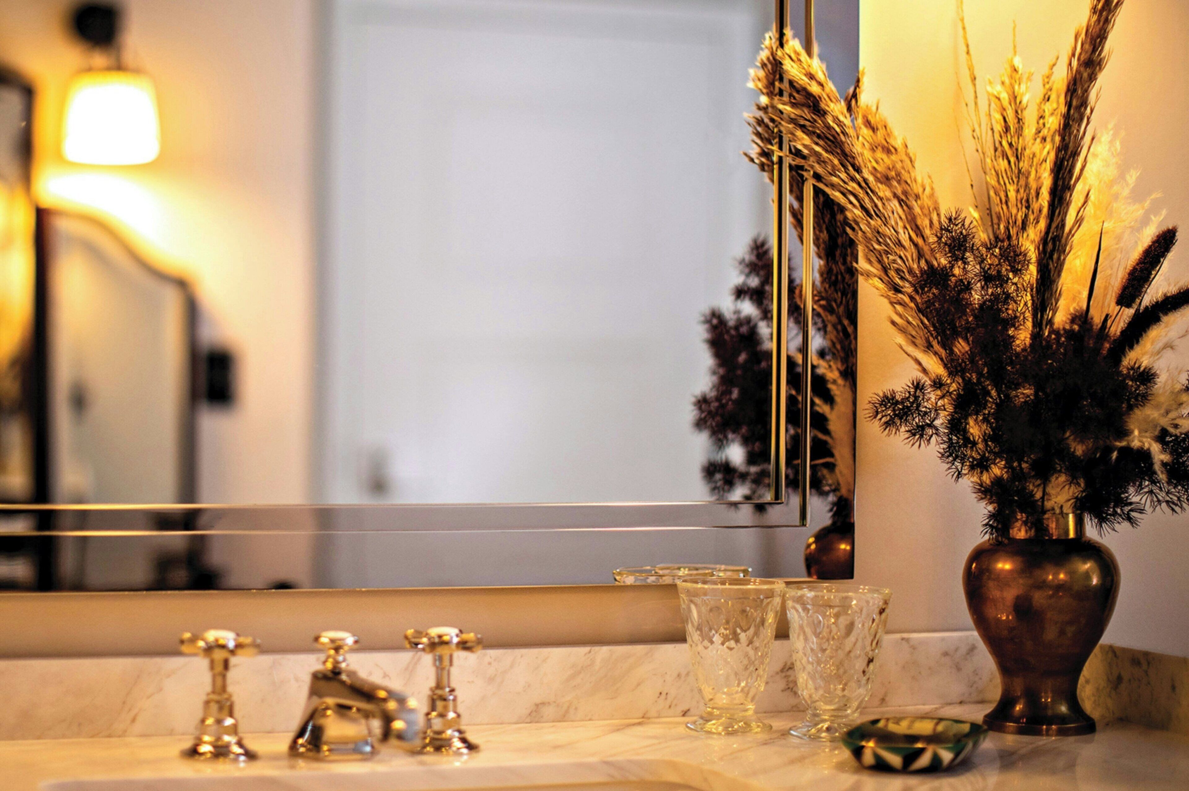 Une salle de bain avec un comptoir en marbre, un robinet doré, un vase en verre contenant des plantes séchées et un miroir qui reflète la pièce.