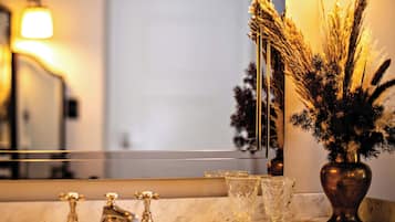 Une salle de bain avec un comptoir en marbre, un robinet doré, un vase en verre contenant des plantes séchées et un miroir qui reflète la pièce.