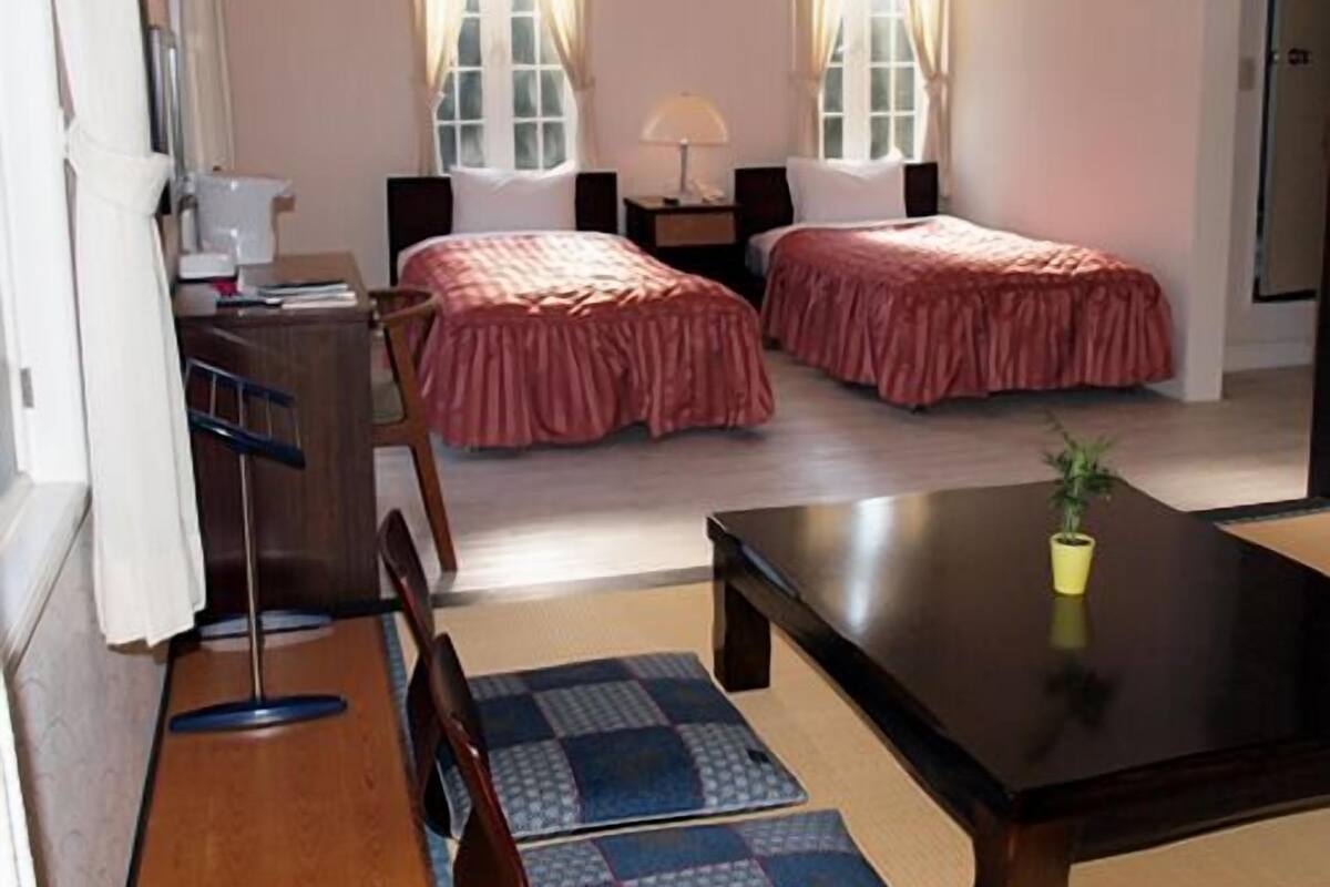 A room with two beds, a desk, a chair, a table, a lamp, and a potted plant.