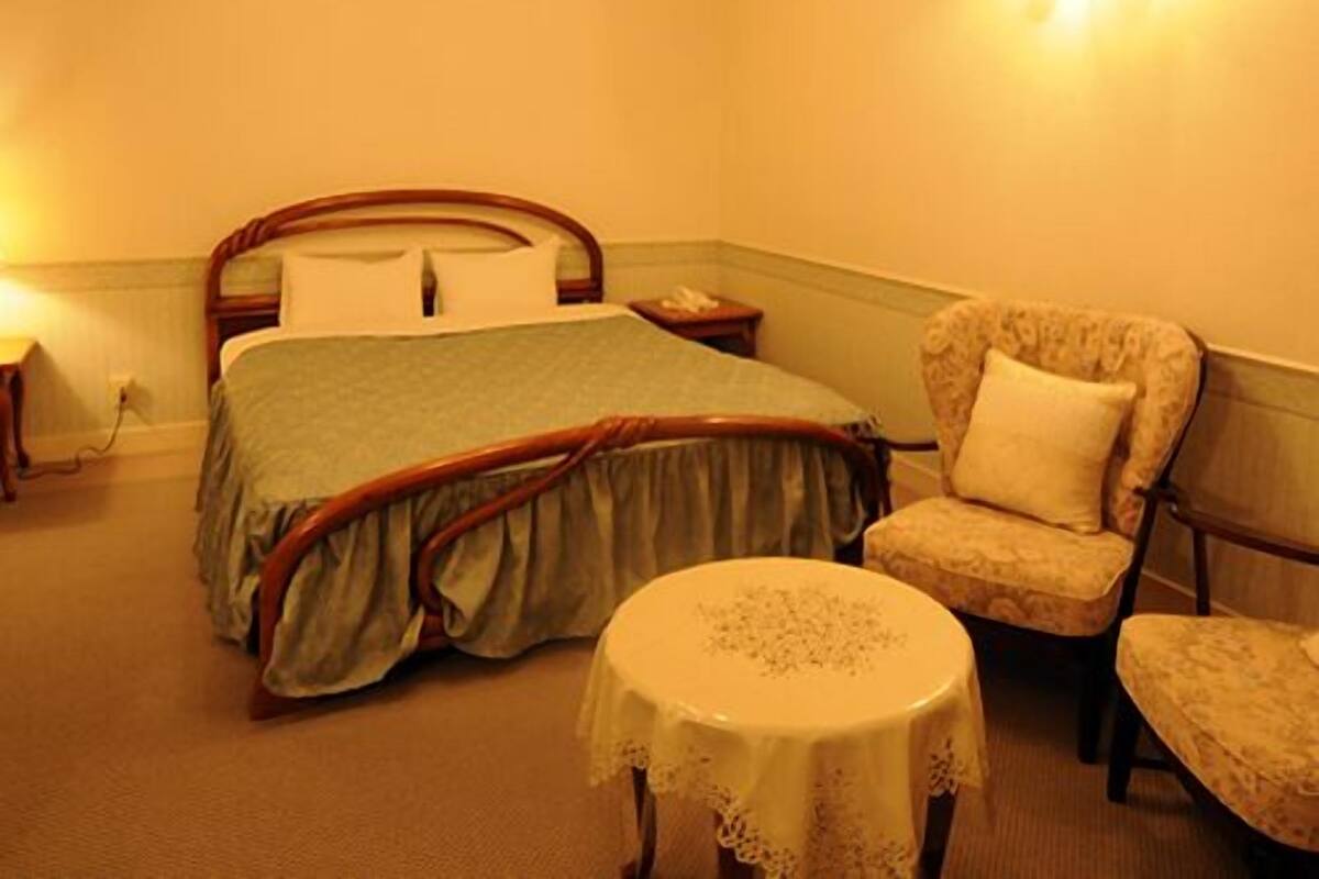 A hotel room with a bed, a chair, a small table, and a wall-mounted light fixture.
