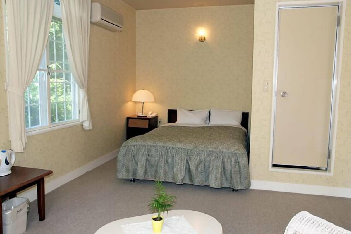 A bedroom with a bed, a bedside lamp, a small table with a plant, a window with curtains, and an air conditioning unit.
