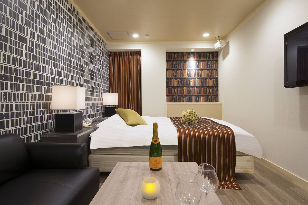 A hotel room with a bed, a bedside table with a bottle of champagne, a lamp, and a bookshelf.