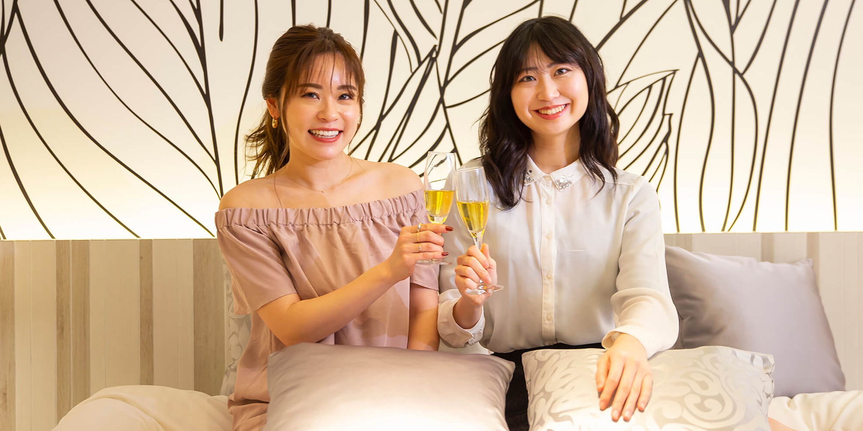 Two individuals seated on a bed with a patterned wall behind them.