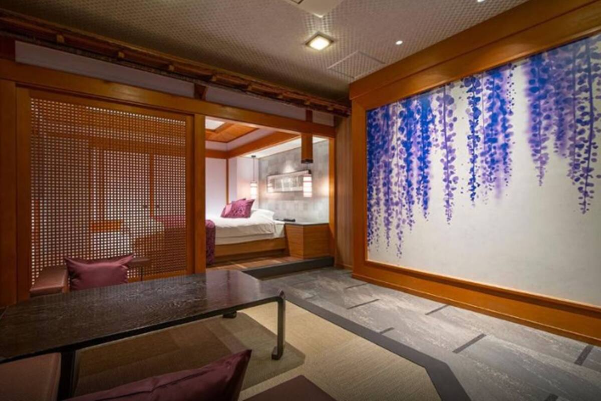 A hotel room with a bed, a desk, a chair, a large wall with a hanging floral design, and a wooden partition.