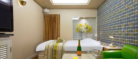 Habitación de hotel con cama, televisión, espejo, silla verde, mesita con una botella y vasos, y un arreglo floral decorativo.