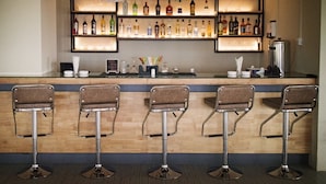 Un espace bar avec un comptoir en bois, quatre tabourets et des étagères remplies de bouteilles d’alcool.