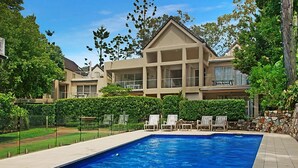 Uma piscina com espreguiçadeiras, cercada por um sebe e um muro de pedra, com uma casa grande ao fundo.