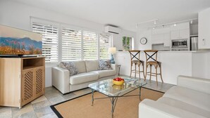 Uma sala de estar moderna com uma mesa de centro de vidro, sofá branco e uma área de cozinha com armários e eletrodomésticos brancos.