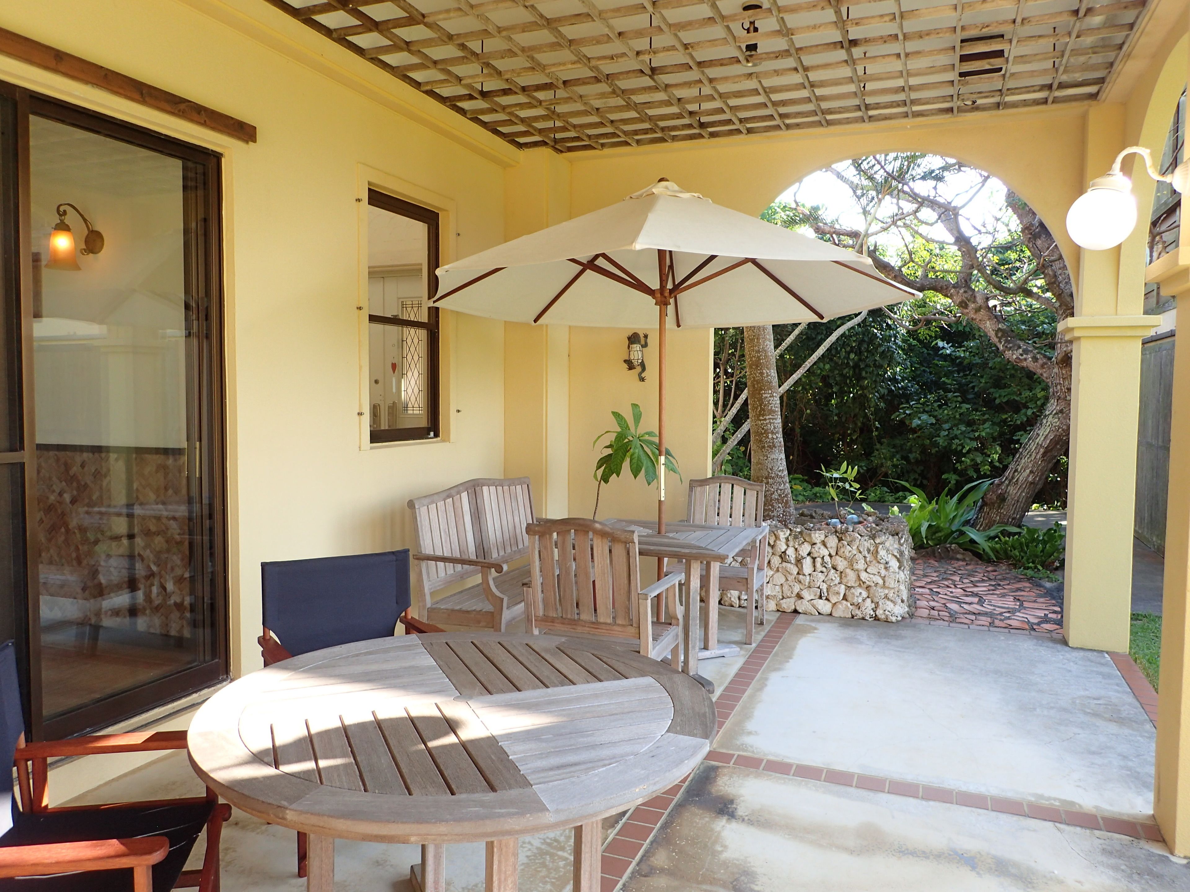 A covered patio area with wooden furniture, including a round table and chairs, and a large umbrella providing shade.