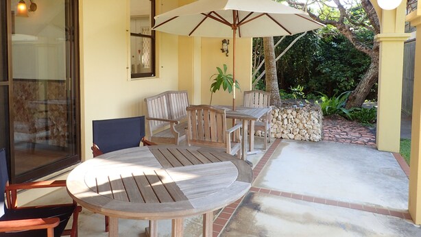 Un patio cubierto con mobiliario de madera, que incluye una mesa redonda y sillas, y un gran paraguas que da sombra.