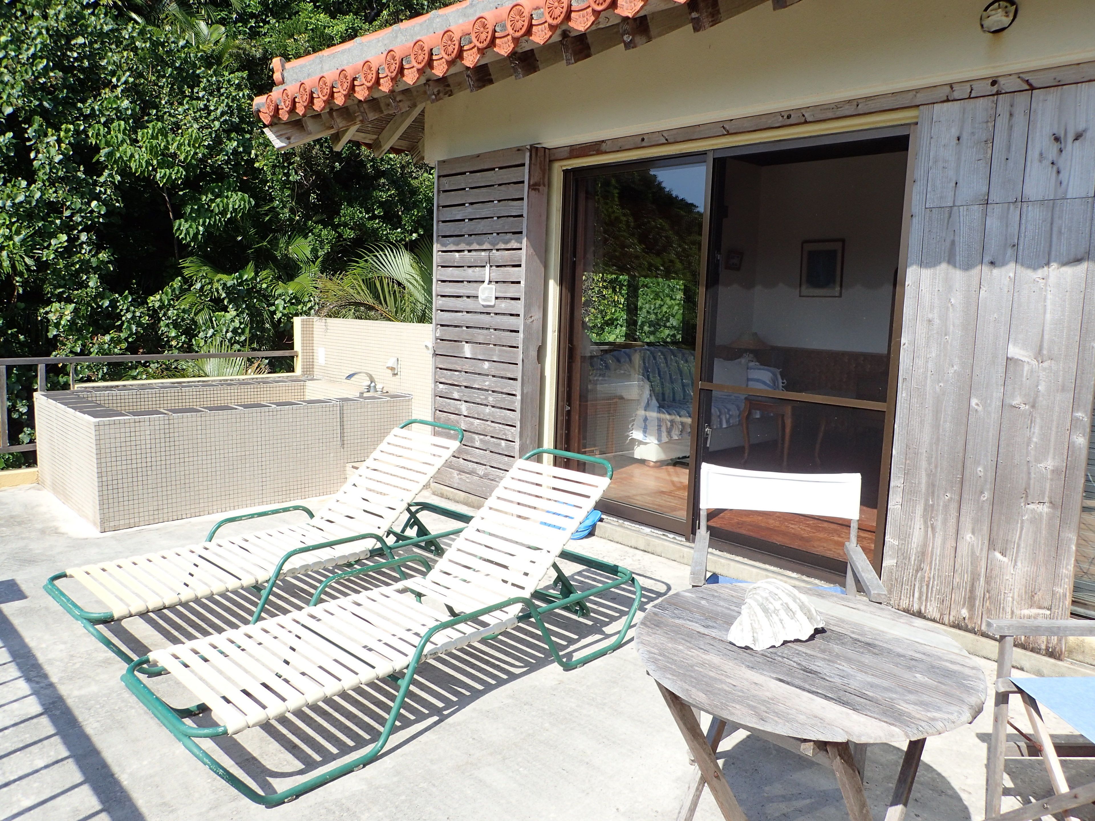 A sunlit outdoor area with two lounge chairs and a table, a tiled pool, and a wooden deck.
