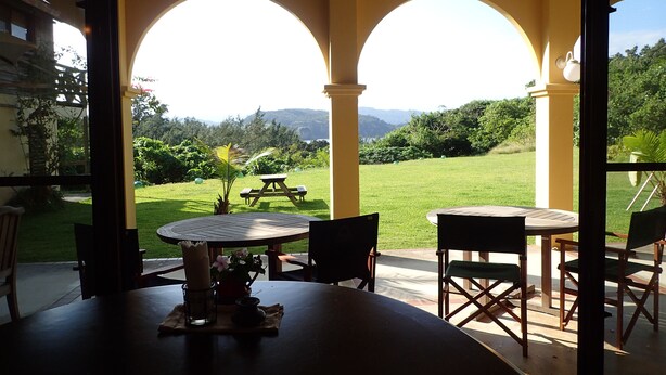 A covered patio with two round tables and chairs, a small table with a candle and flowers, and a view of a grassy area with trees and mountains in the distance. - MaYaGuSuKu RESORT (Taketomi)
