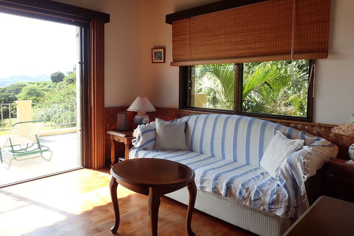 A room with a sofa, a small table, a lamp, and a window with a view of greenery.