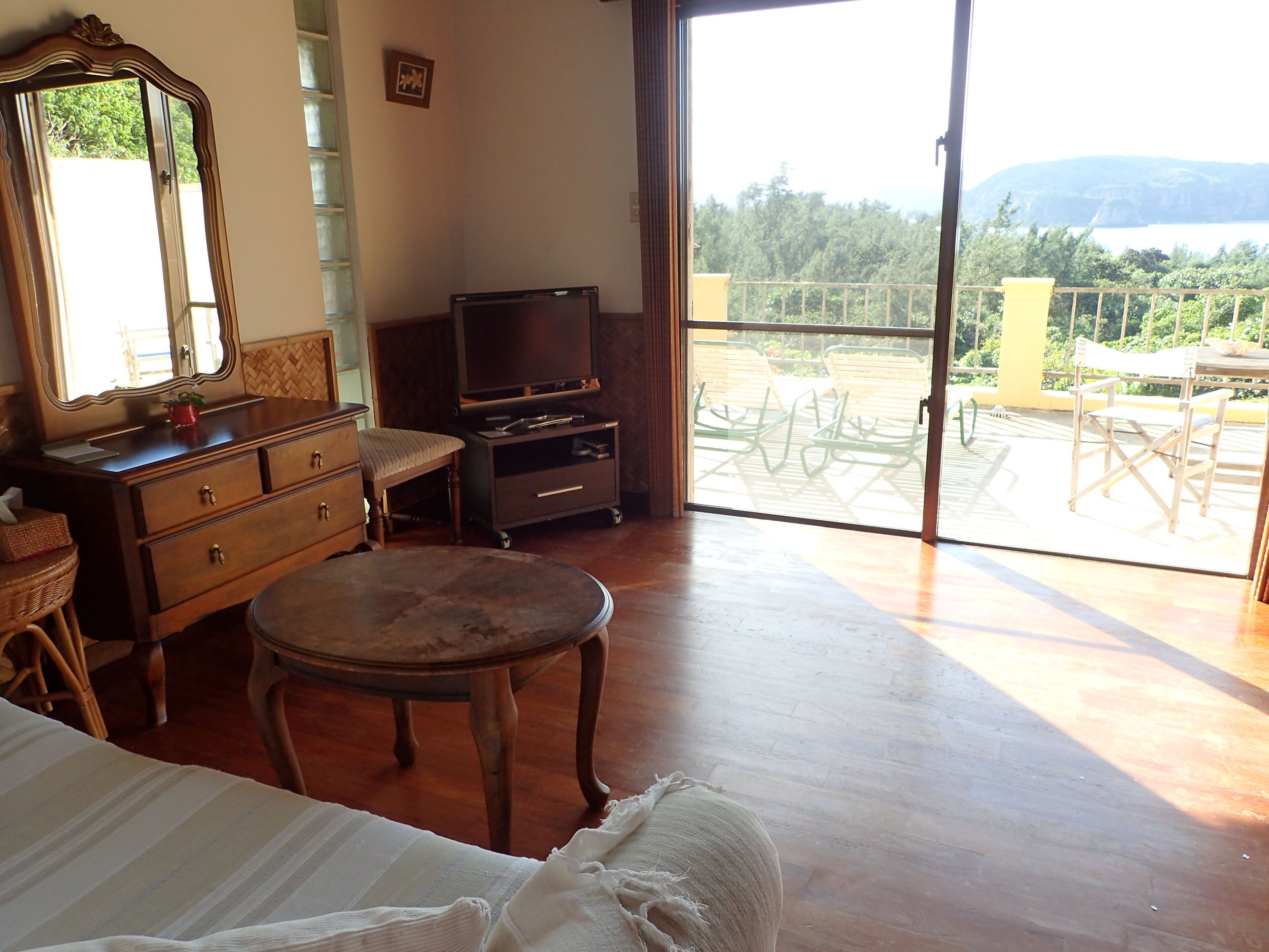 A room with a bed, a dresser, a television, and a balcony with outdoor furniture.