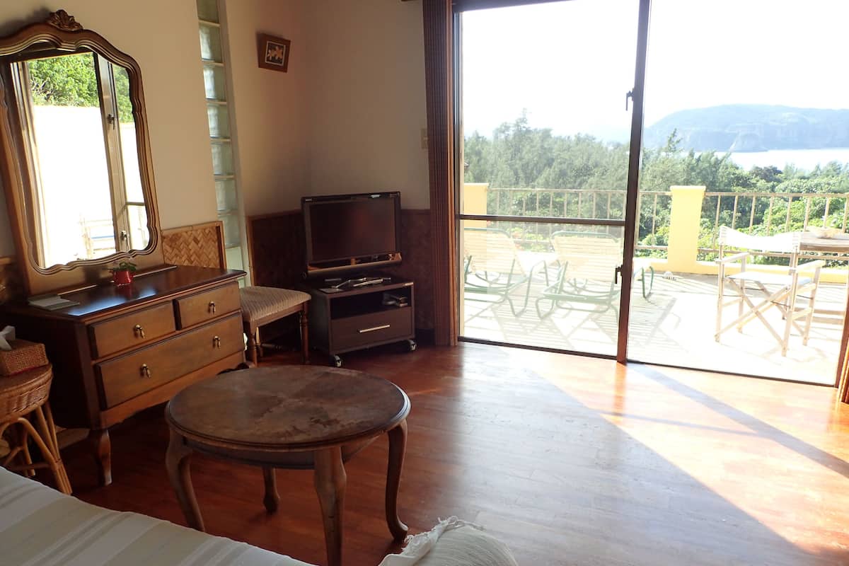 A room with a bed, a dresser, a television, and a balcony with outdoor furniture.