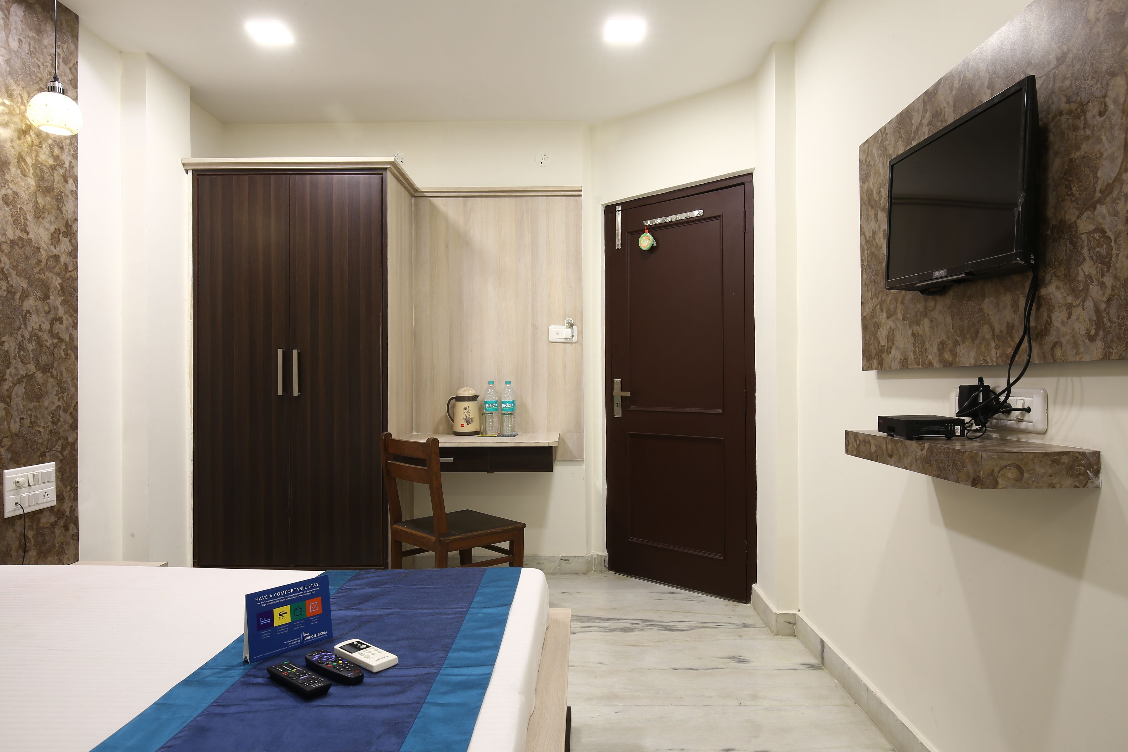 A hotel room with a bed, a desk with a chair, a wardrobe, a television mounted on the wall, and a shelf with a TV and other items.