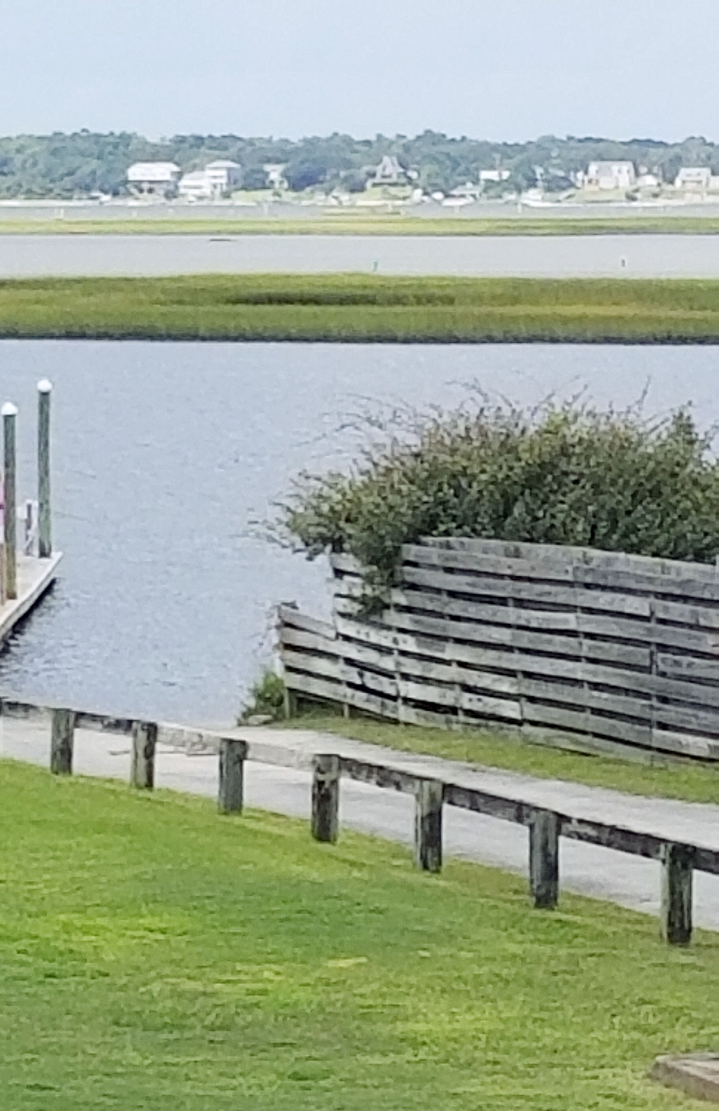 Blick auf das Wasser mit einer Holzsteganlage, grünen Flächen und einer entfernten Küstenlinie mit Gebäuden.