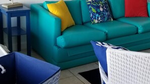 A living room with a teal sofa, a blue and white wicker chair, a blue storage box, and a patterned wallpaper. - Popular   ~ Quaint. Bogue Shores Condo --  just steps to a beautiful beach (Atlantic Beach)