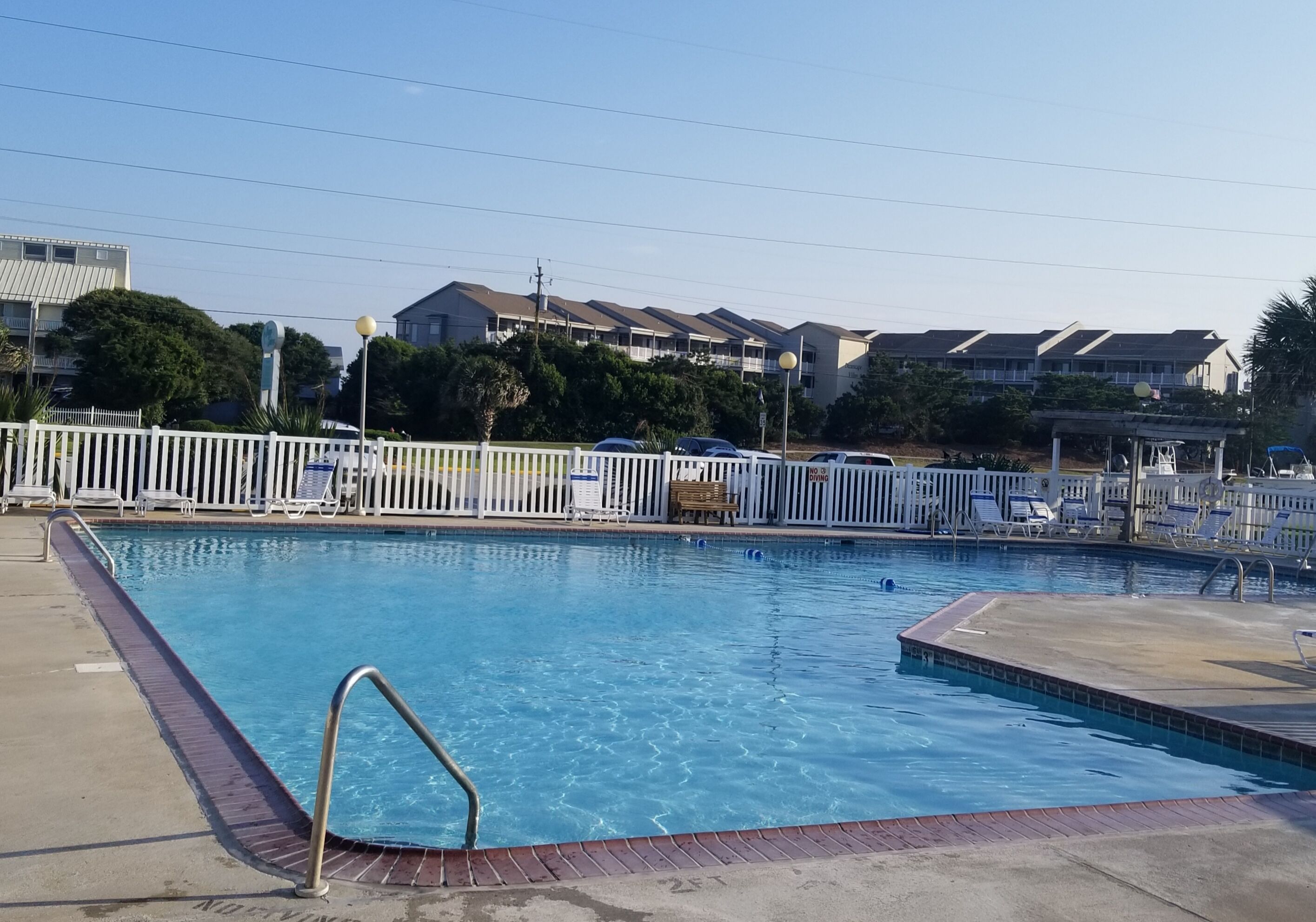 A swimming pool with a white fence, lounge chairs, and a clear sky.
