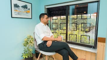 A man sitting by a window with a view of greenery, a framed picture on the wall, and a potted plant beside him.