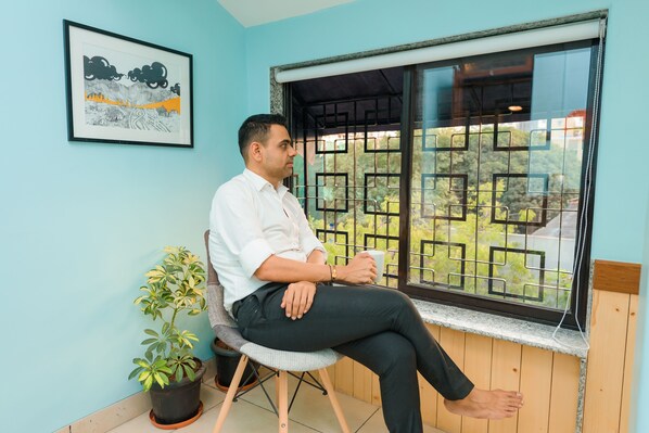 Um homem sentado perto de uma janela com vista para o verde, um quadro na parede e um vaso de planta ao lado dele.
