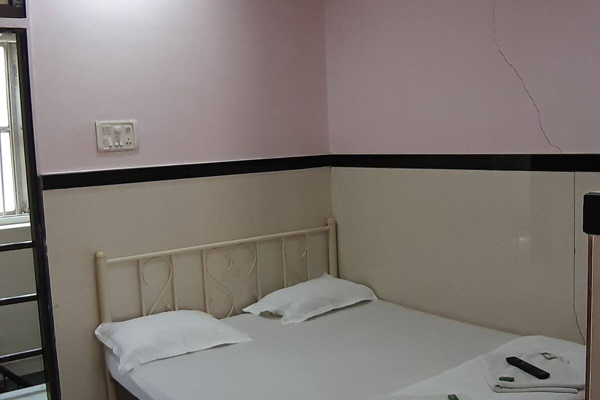 Un lit simple avec du linge de lit blanc, un ventilateur de plafond, une table de chevet et une chaise dans une pièce aux murs roses et aux moulures noires.