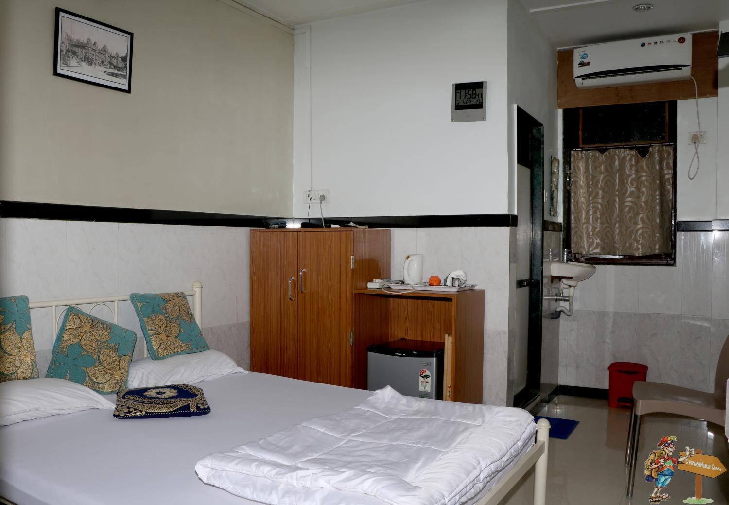 A hotel room with a bed, a wardrobe, a desk with a kettle, a small trash bin, and a bathroom with a sink and shower curtain.