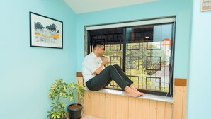 Una persona asseguda en un davantà de finestra d'una habitació amb parets blaves clares, un quadre al mur i dues plantes en testos.
