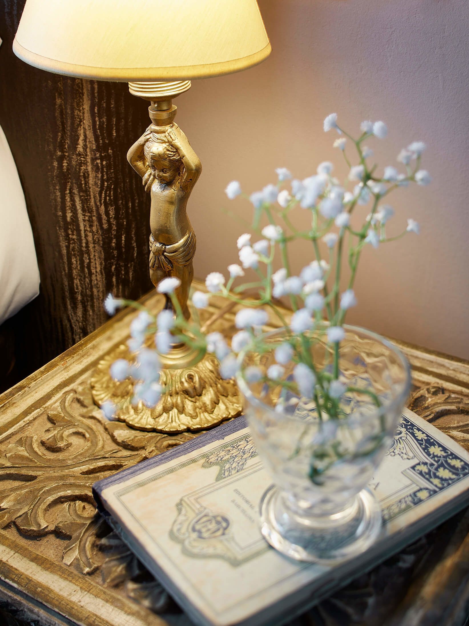 Ein Nachttisch mit einer dekorativen Lampe, einer Glasvase mit Blumen und einem Buch.