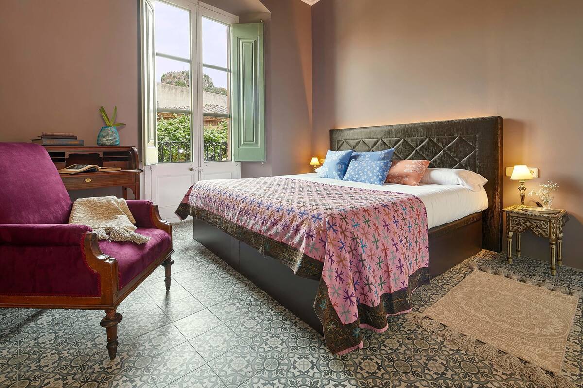 Un dormitorio con cama, una silla, una mesita de noche y una ventana con contraventanas.