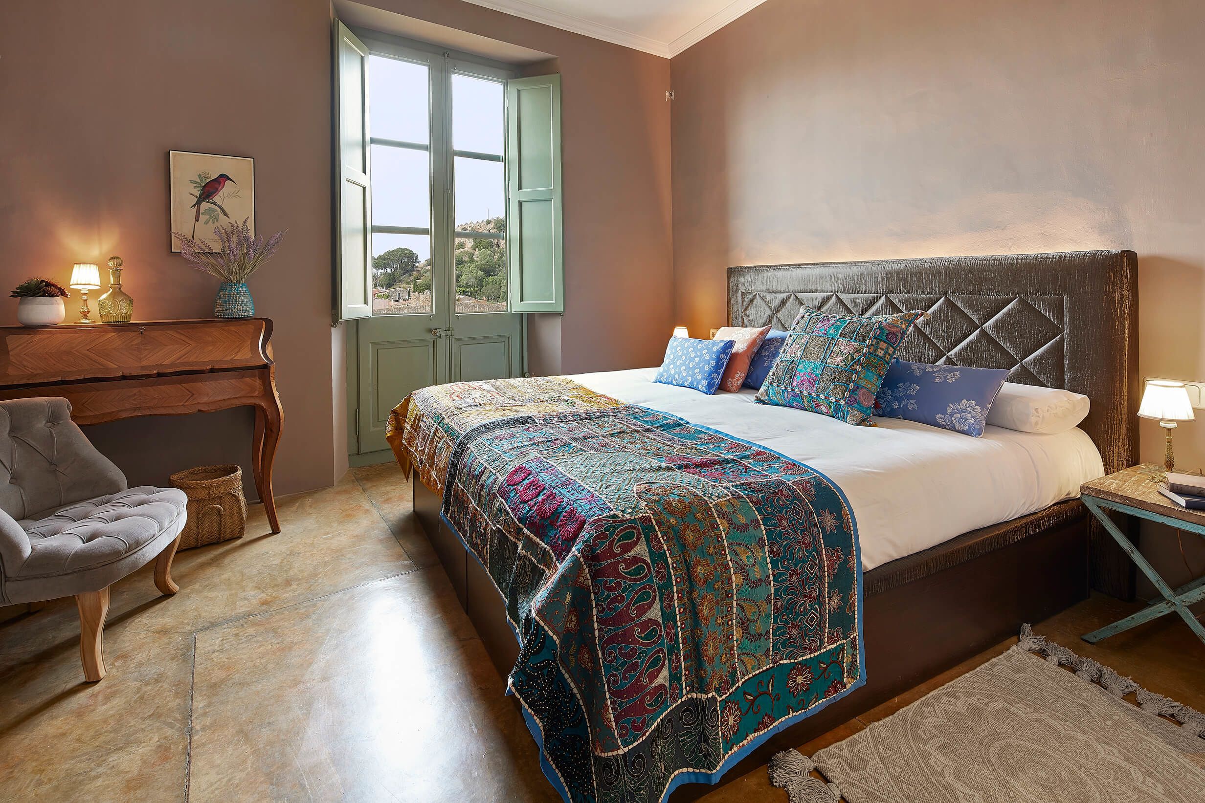 A bedroom with a large bed, a wooden bench, a chair, a window with a view, and a wall-mounted lamp.