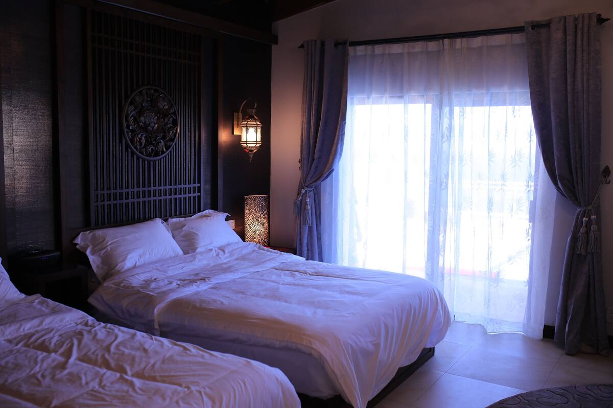 A hotel room with two beds, a decorative wall panel, a wall-mounted lantern, and a large window with curtains.