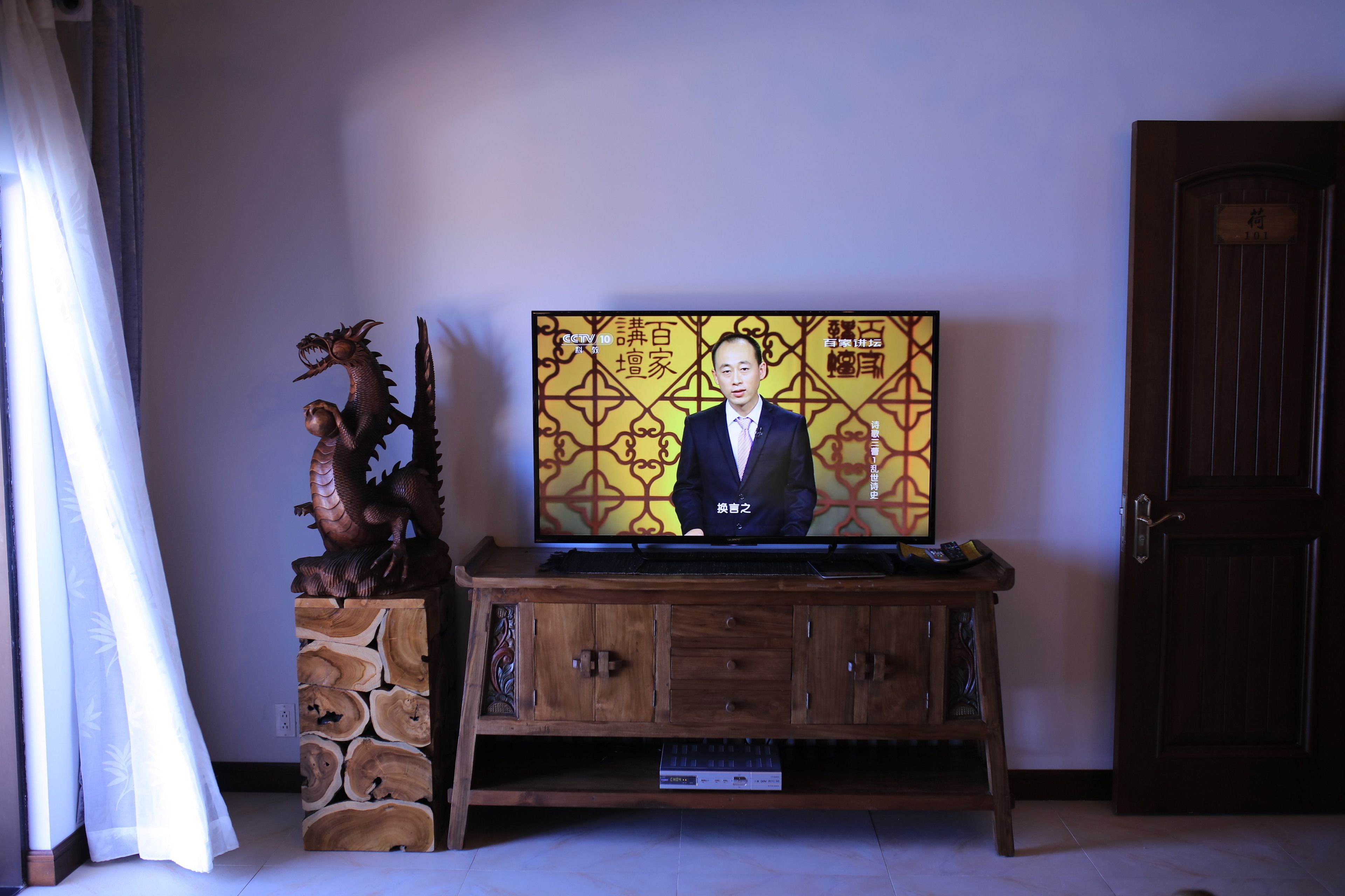 A television on a wooden stand with a dragon figure beside it, a wooden cabinet below, and a door in the background.