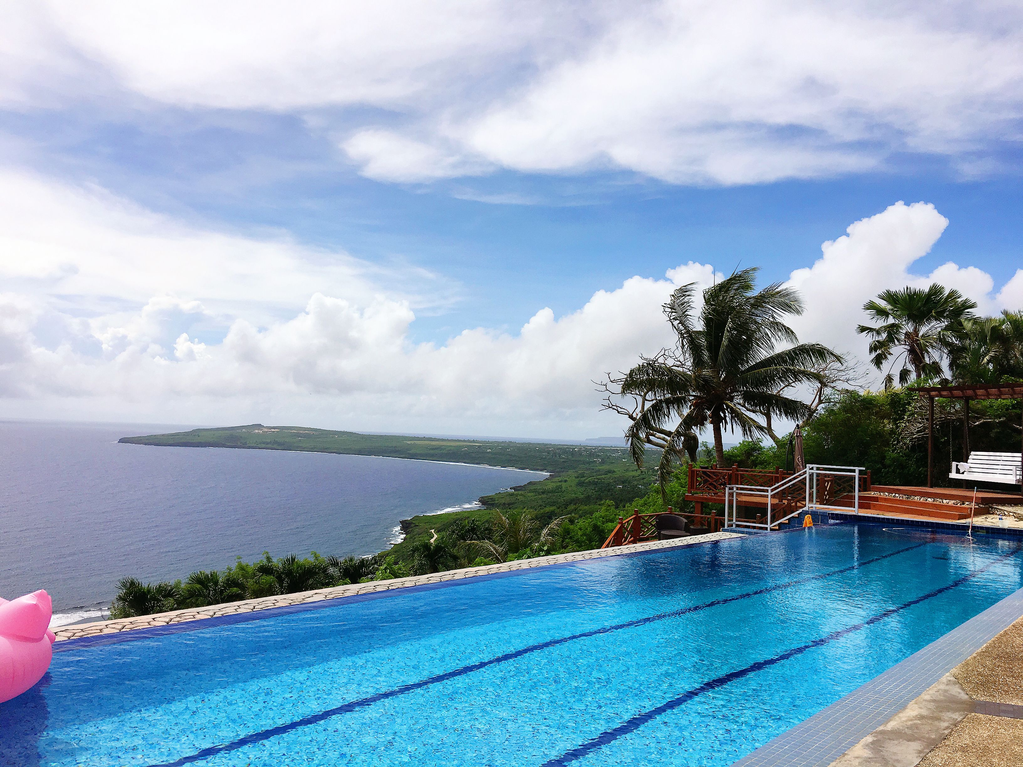A swimming pool with clear blue water, a wooden deck, and a view of the ocean and hills in the background.