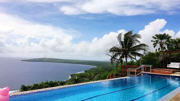A swimming pool with clear blue water, a wooden deck, and a view of the ocean and hills in the background.