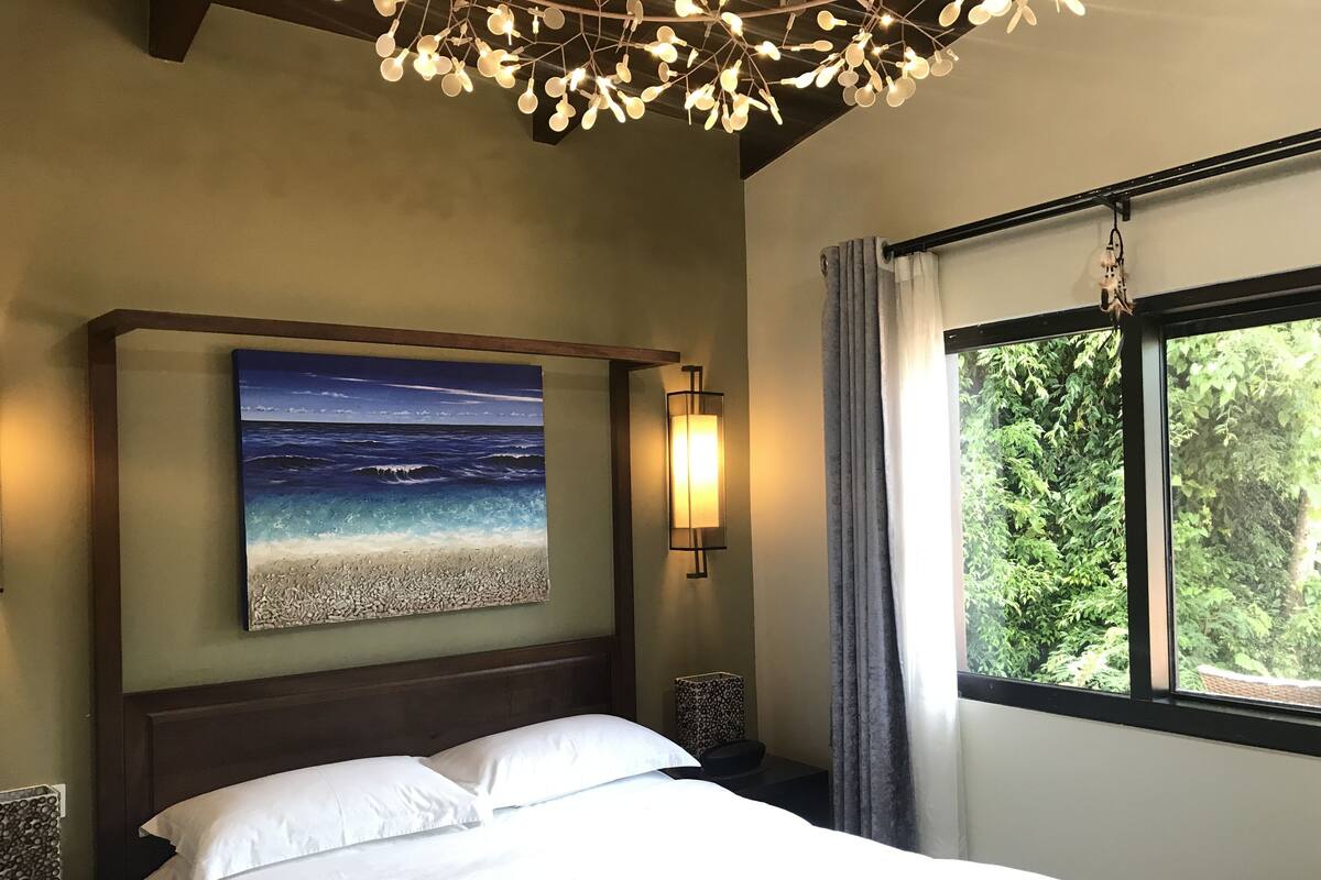 A bedroom with a bed, a painting of a beach scene, a wall-mounted light, and a window with curtains.