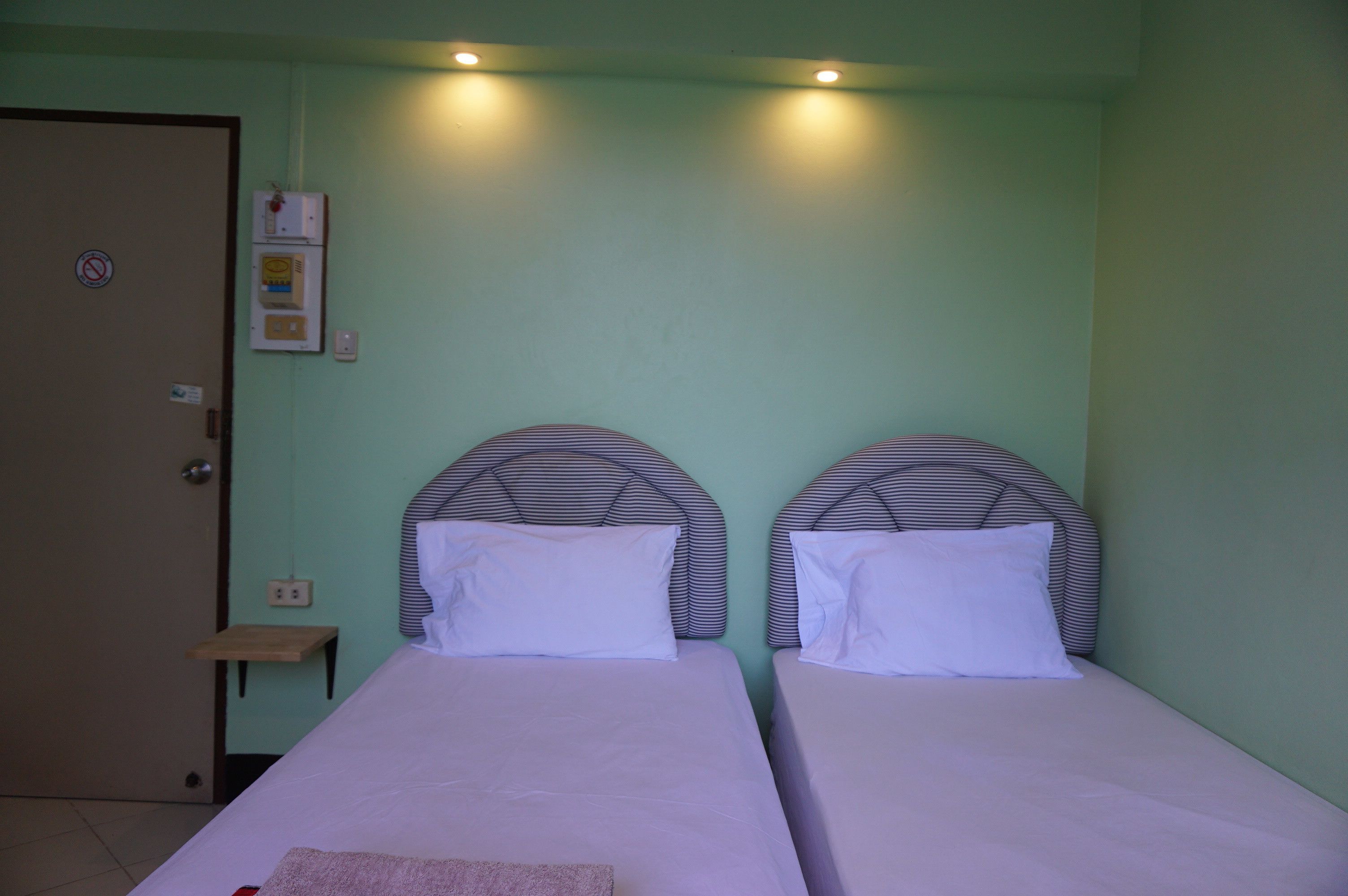A room with two beds, a wooden nightstand, a wall-mounted electrical outlet, and a door with a 'no smoking' sign.
