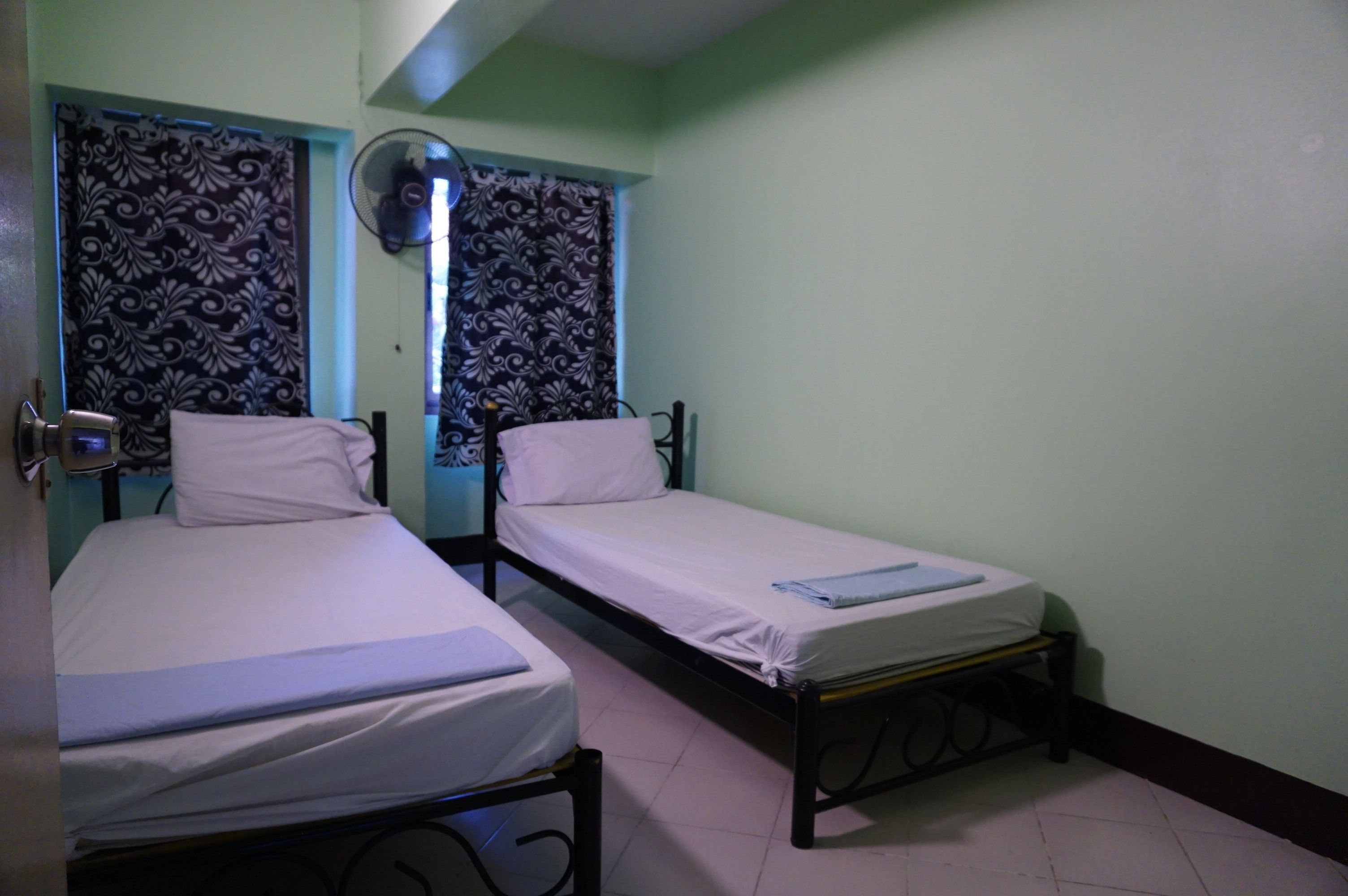 Two single beds with white linens, a wall-mounted fan, and patterned curtains.