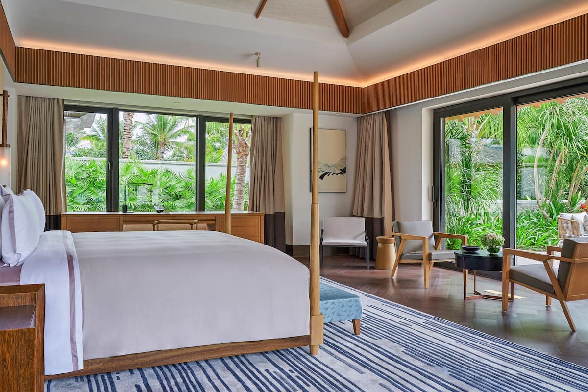 Une chambre d’hôtel moderne, dotée d’un grand lit, d’un bureau et d’une chaise. De grandes fenêtres offrent une vue sur la verdure environnante.