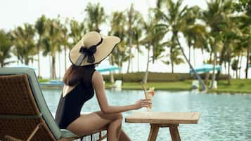 A person relaxing by a poolside lounge chair with a drink, surrounded by palm trees and a clear view of the water.