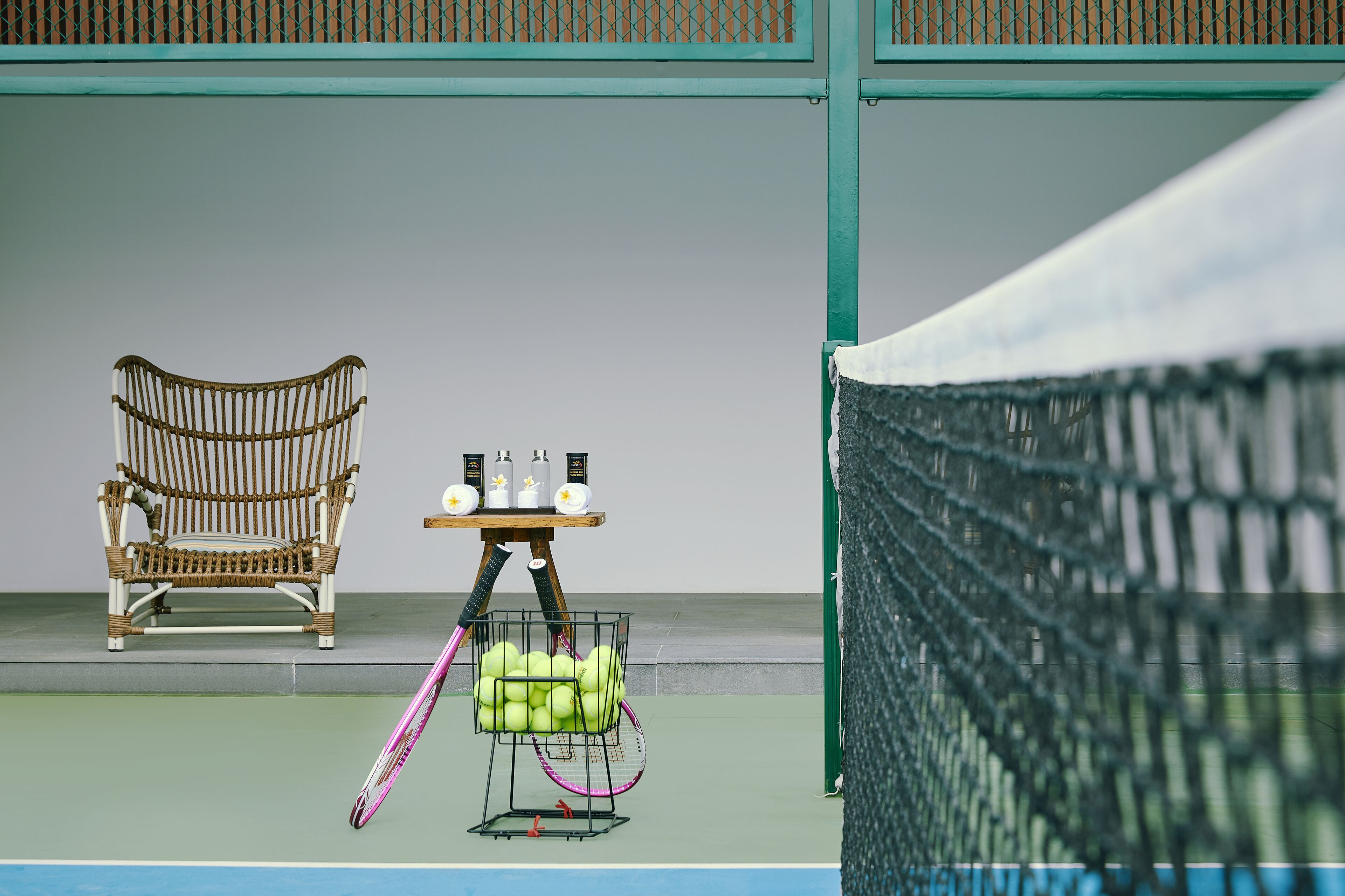 Una cancha de tenis con una silla de mimbre, una mesa con botellas y una raqueta de tenis.