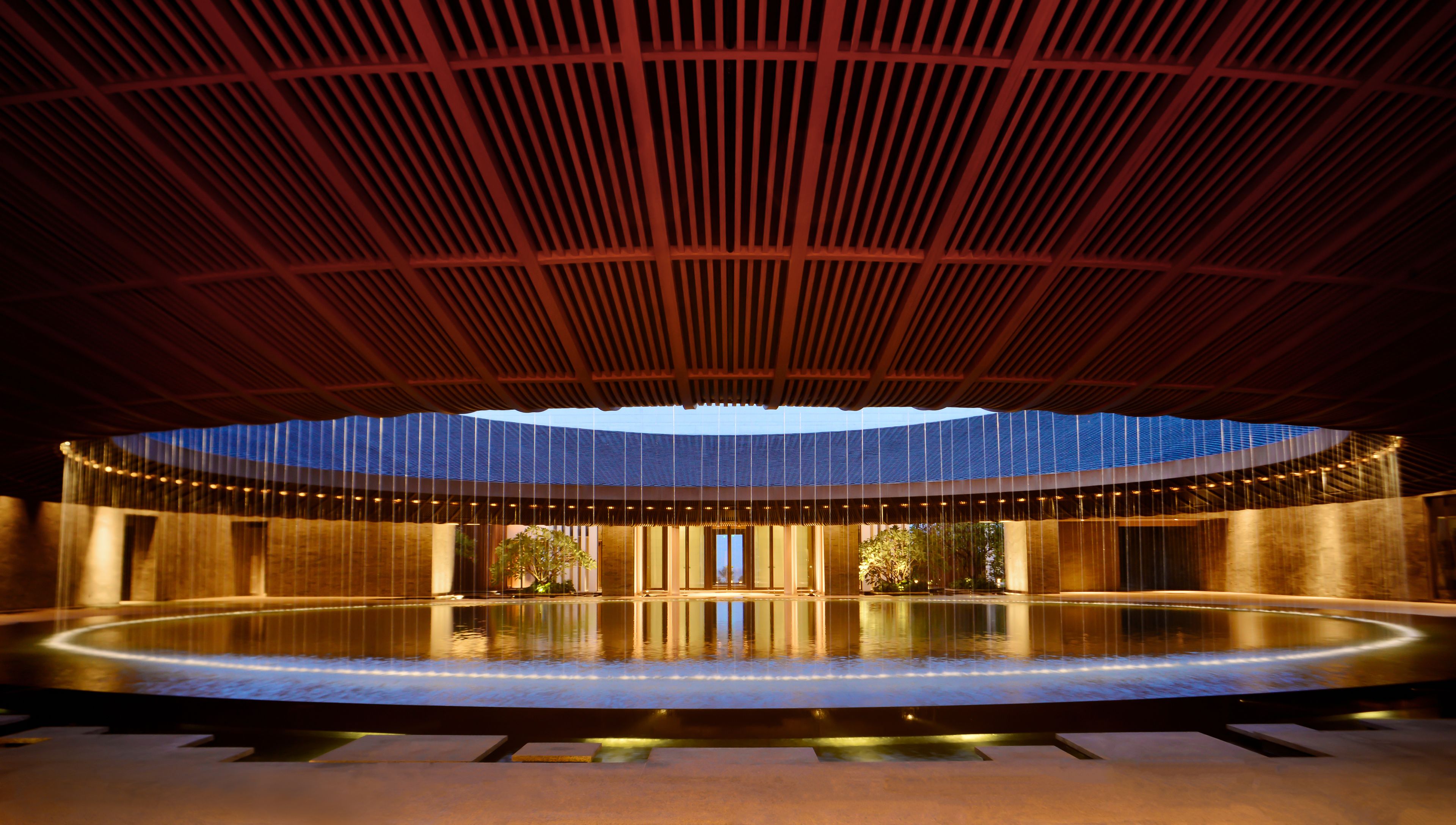 A spacious indoor area with a large circular water feature, a high ceiling with a wooden slatted design, and a view of the sky through a large opening.