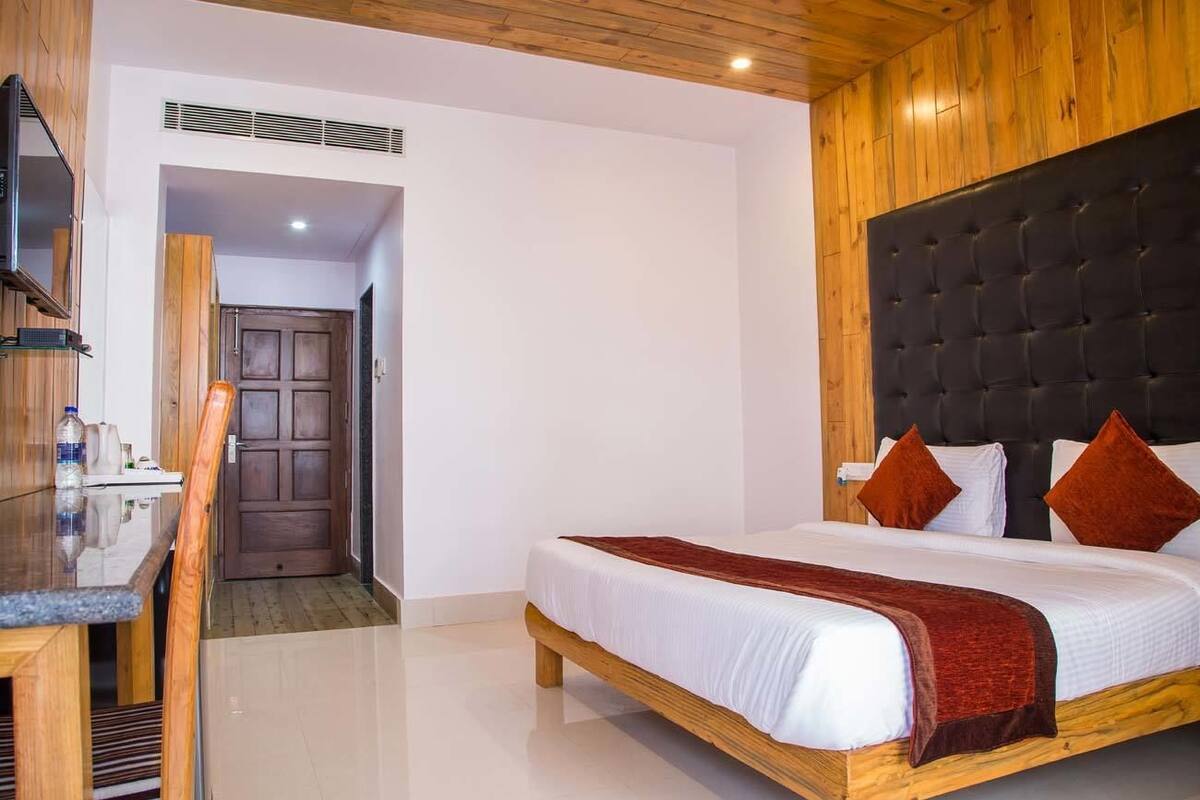 A bedroom with a wooden headboard, a bed with white linens and two brown pillows, a wooden desk with a chair, and a wooden door in the background.
