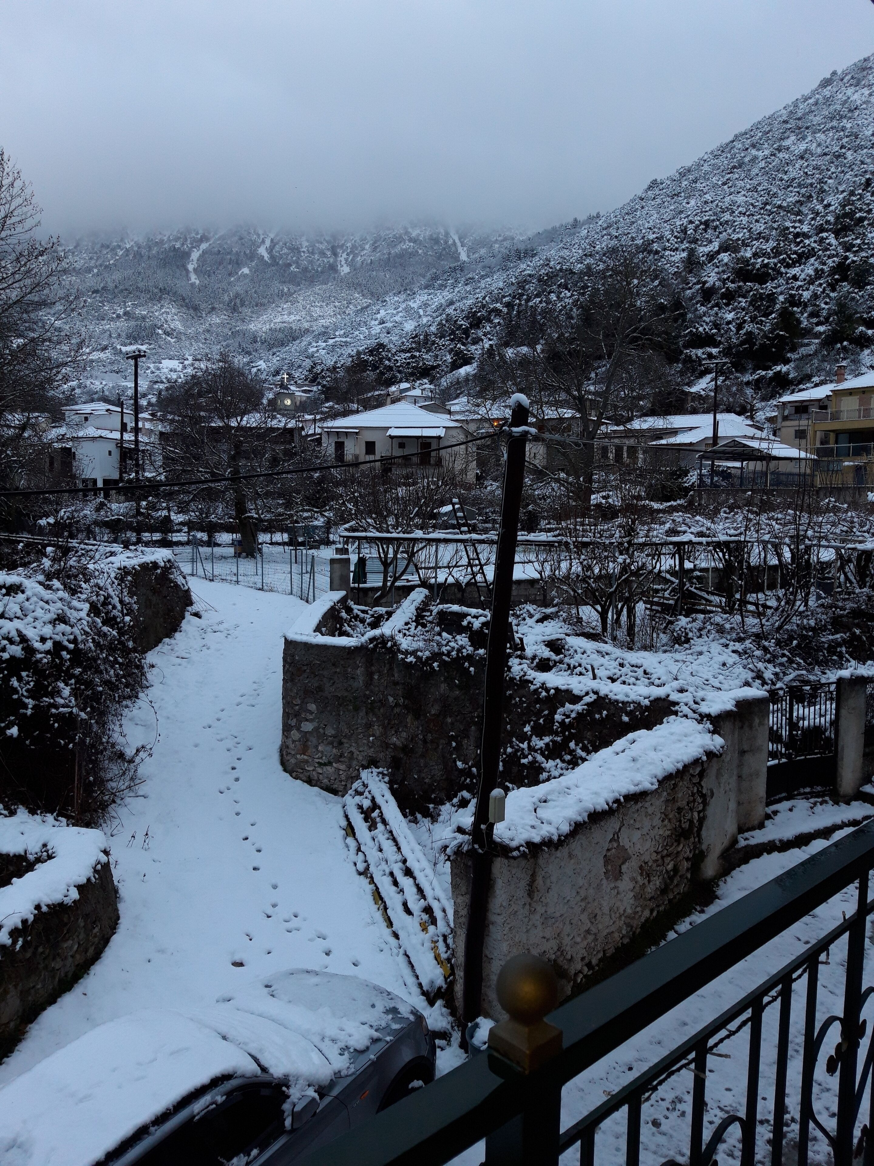 A snow-covered village with traditional houses, a stone wall, and a car parked in the foreground.