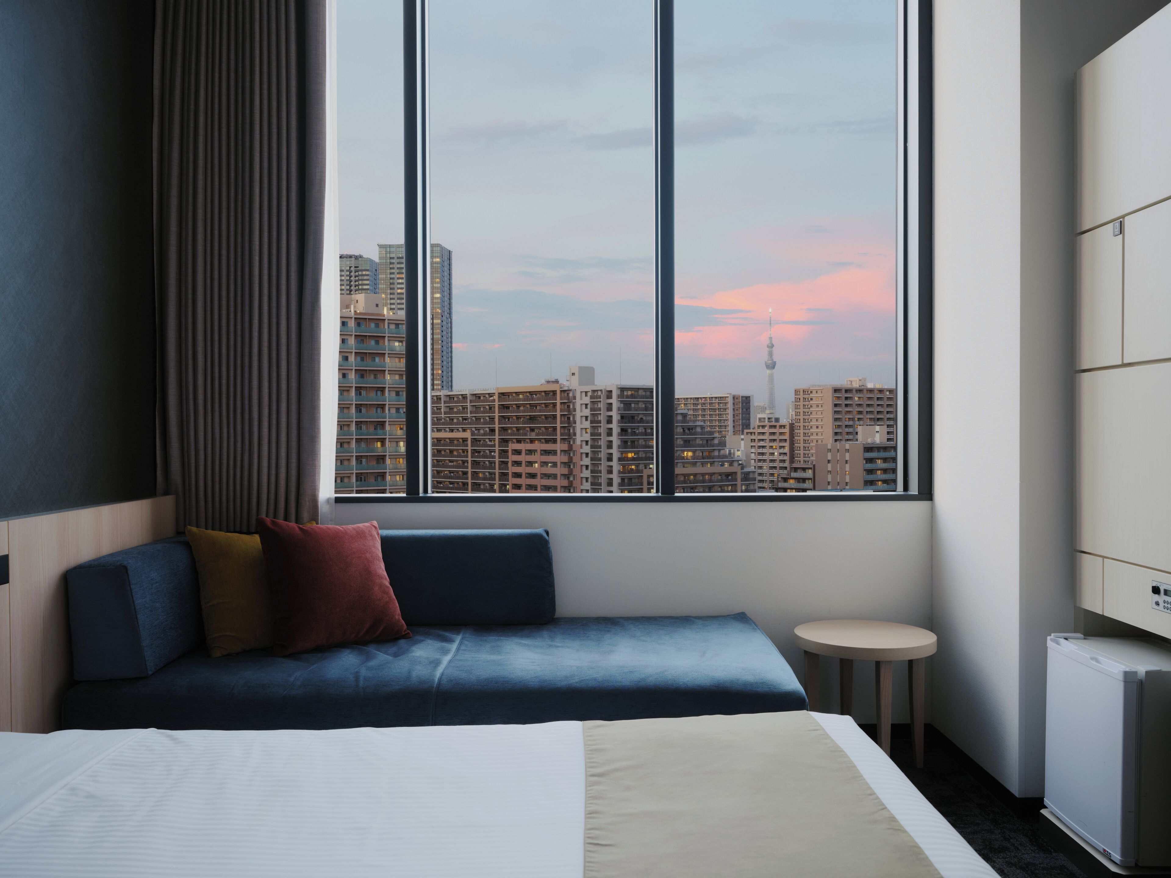 Ein Hotelzimmer mit großem Fenster, einem Bett, einer Sitzecke mit Kissen, einem kleinen runden Tisch und einem Heizkörper.