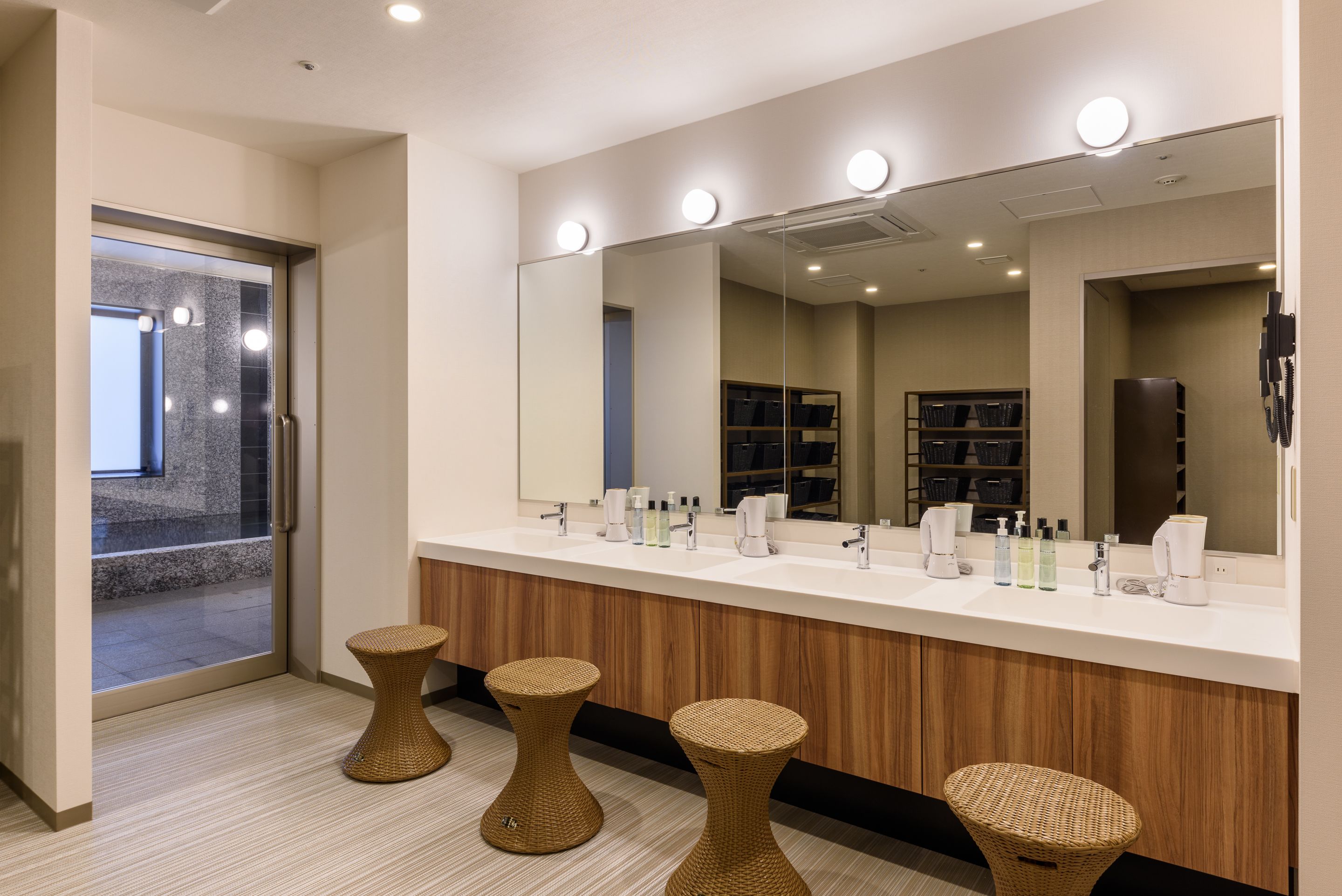 A modern bathroom with a large mirror, two stools, and a vanity with multiple sinks and toiletries.