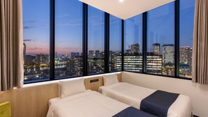 Ein Hotelzimmer mit zwei Betten, Blick auf eine Stadtlandschaft im Abendlicht und einem großen Fenster mit Vorhängen.